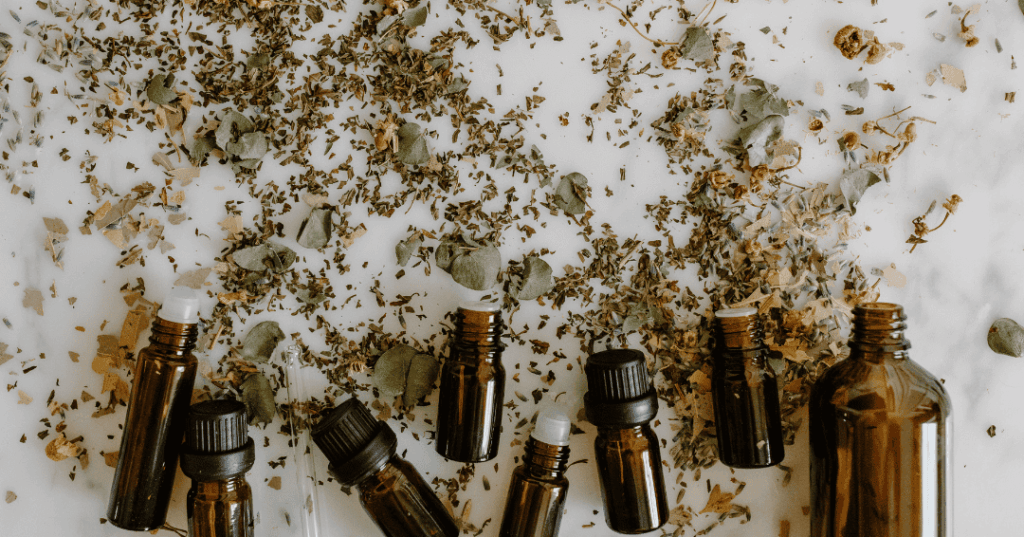 Diverse braune Glasflaschen mit ätherischen Ölen liegen auf einem weißen Untergrund, darüber sind viele Kräuter verteilt.